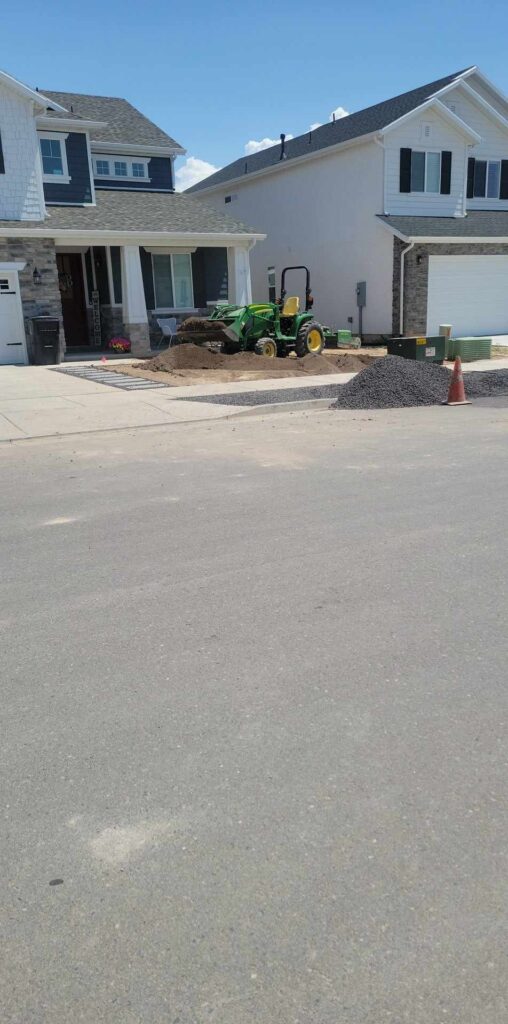 Picture of a house across the street, with a green tractor in a dirt lawn and a large pile of pebbles