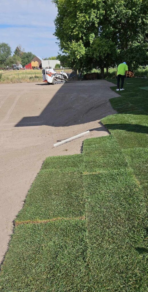 A large open space with smooth dirt, a small white tractor and single worker on the side laying sod.