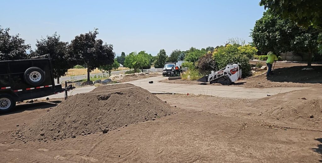 Large yard with a trailer, small tractor and truck. Large pile of dirt and one person working under a tree