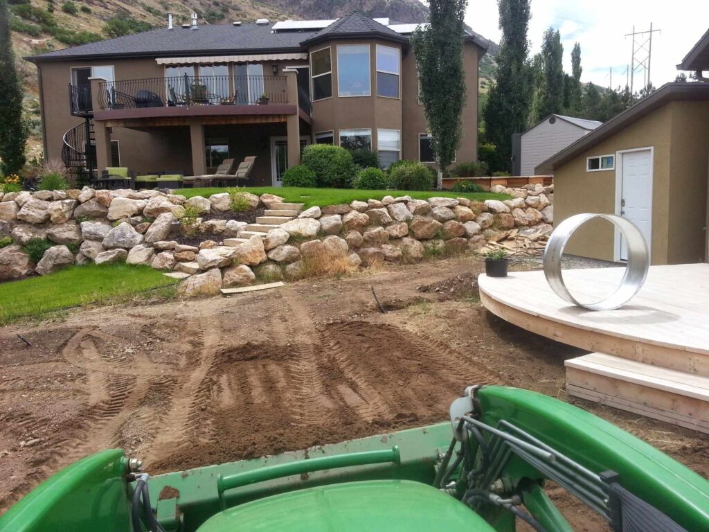 Picture taken from seat of green tractor, smooth dirt, small boulders, stair case, wooden patio with metal ring, and two buildings.