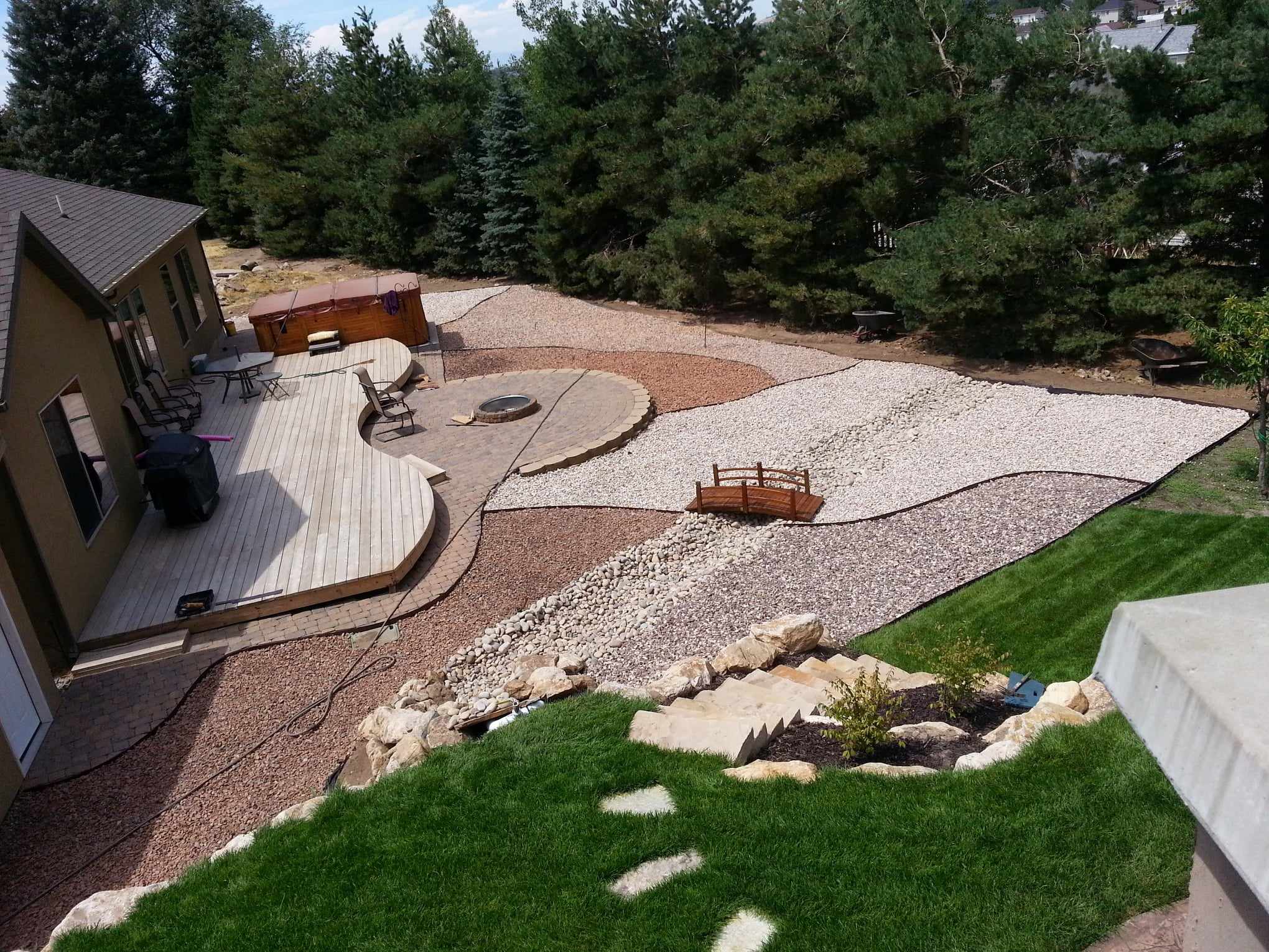 Picture overlooking grassy area, and a large open space surrounded by a building and trees on the opposite side. A wooden patio is pictured. Large open rock landscaping with three colors of rock, a streambed, small bridge, and firepit